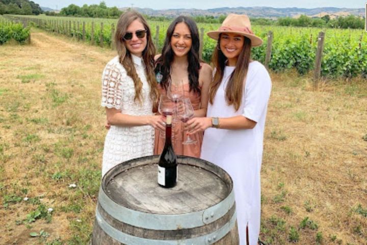 Three women holding wine glasses stand by a barrel in a vineyard.