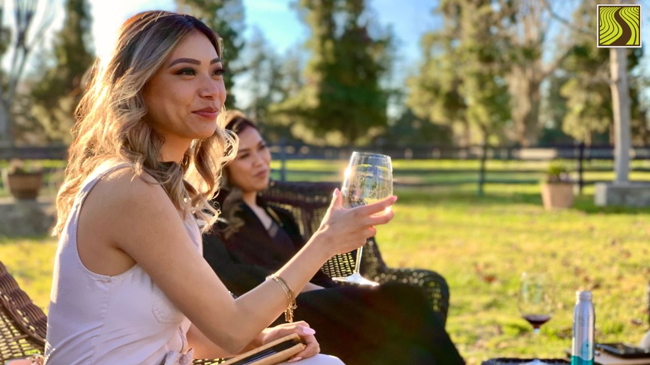 Two women sitting outdoors, holding wine glasses, with a sunny garden in the background.