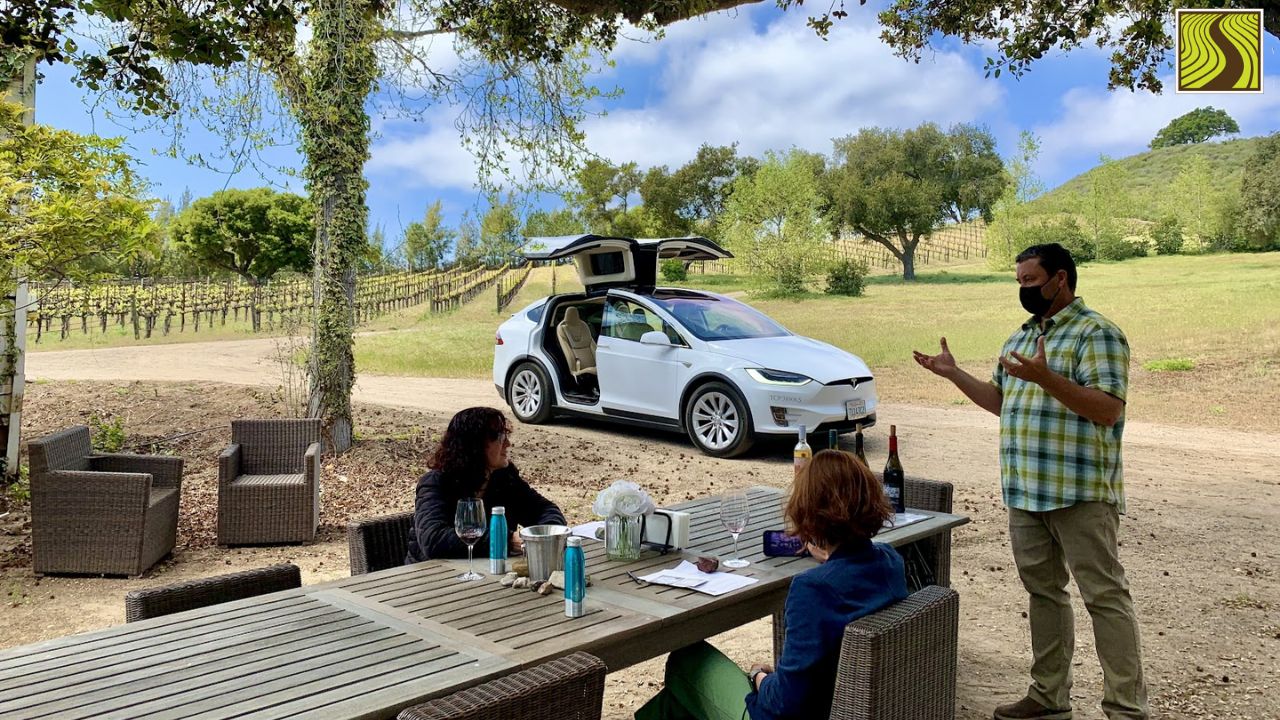 People tasting wine outdoors by a table, with an electric car parked nearby.