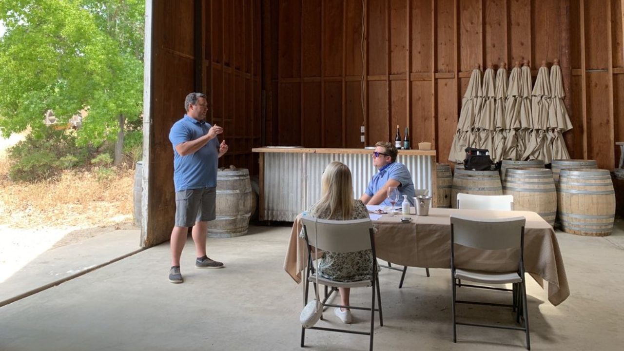 Person standing near a table with two seated individuals in a barn with wine barrels.
