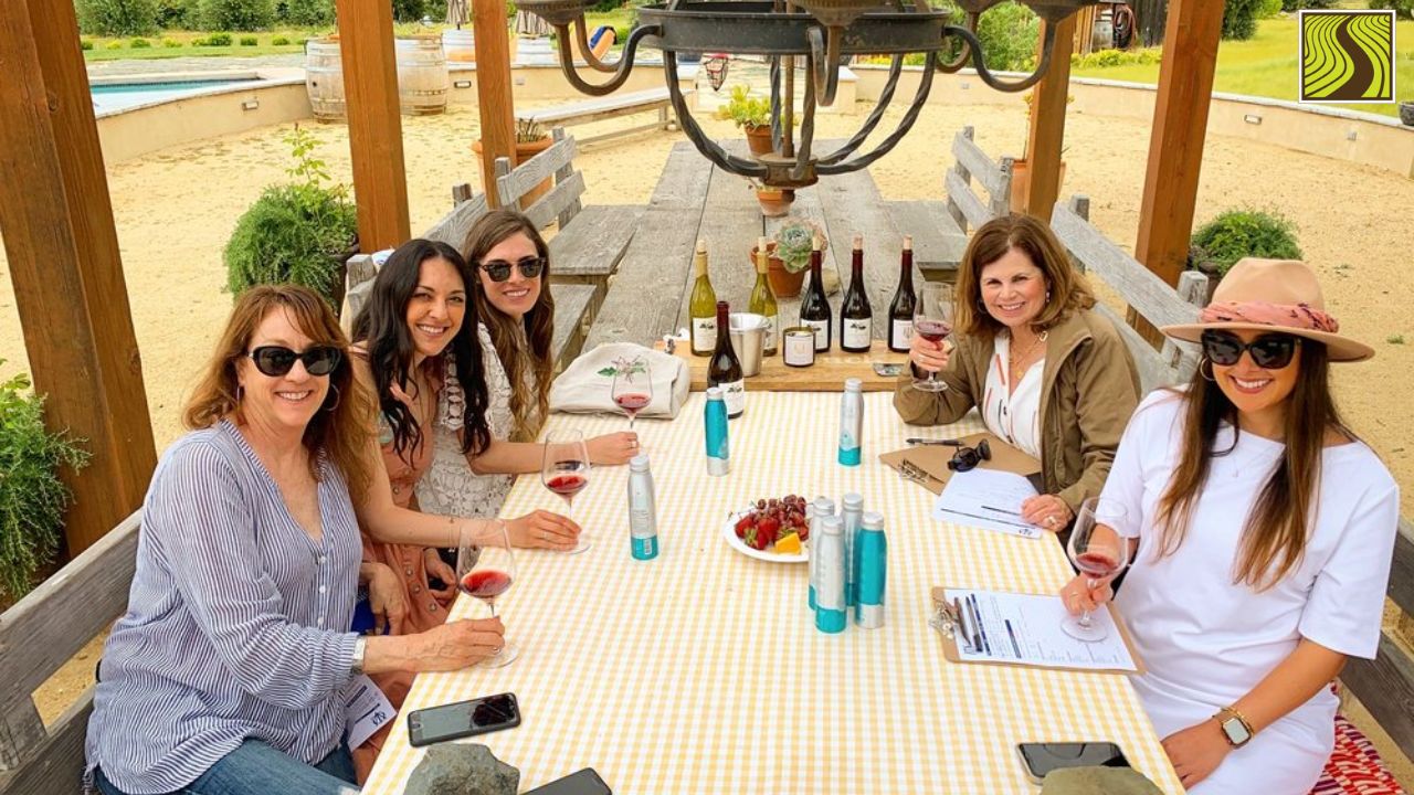 Five women sitting at a table outdoors, holding wine glasses and smiling.
