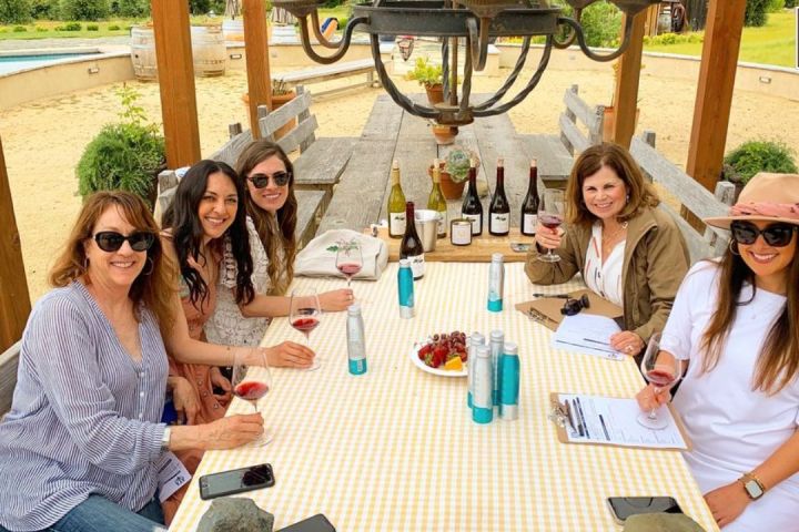 Five women sitting at a table outdoors, holding wine glasses and smiling.