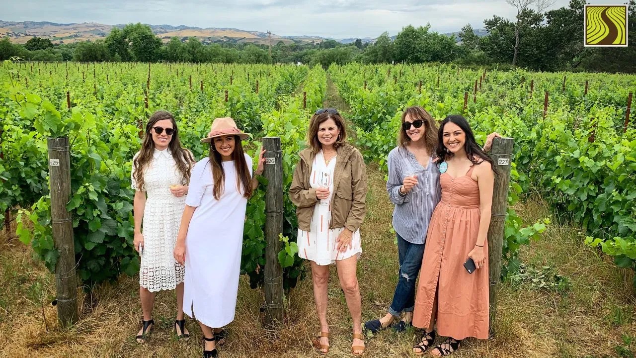 Five women standing in a vineyard, posing and smiling for the camera.