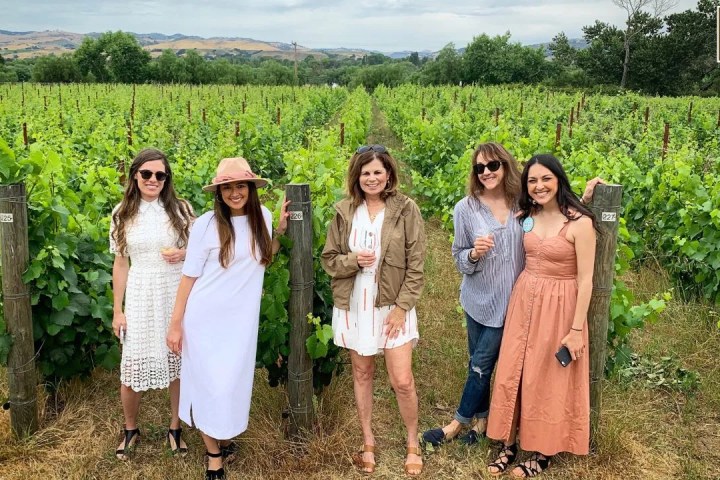 Five women standing in a vineyard, posing and smiling for the camera.