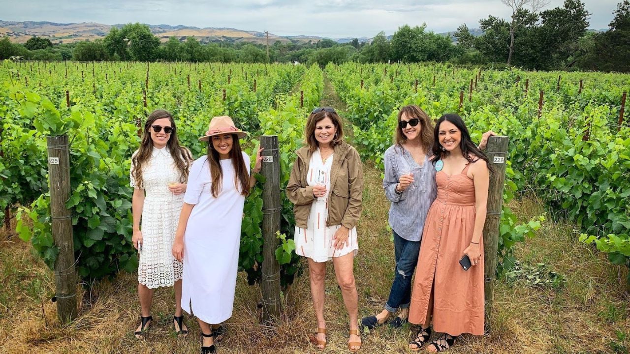 Five women standing in a vineyard, smiling at the camera.